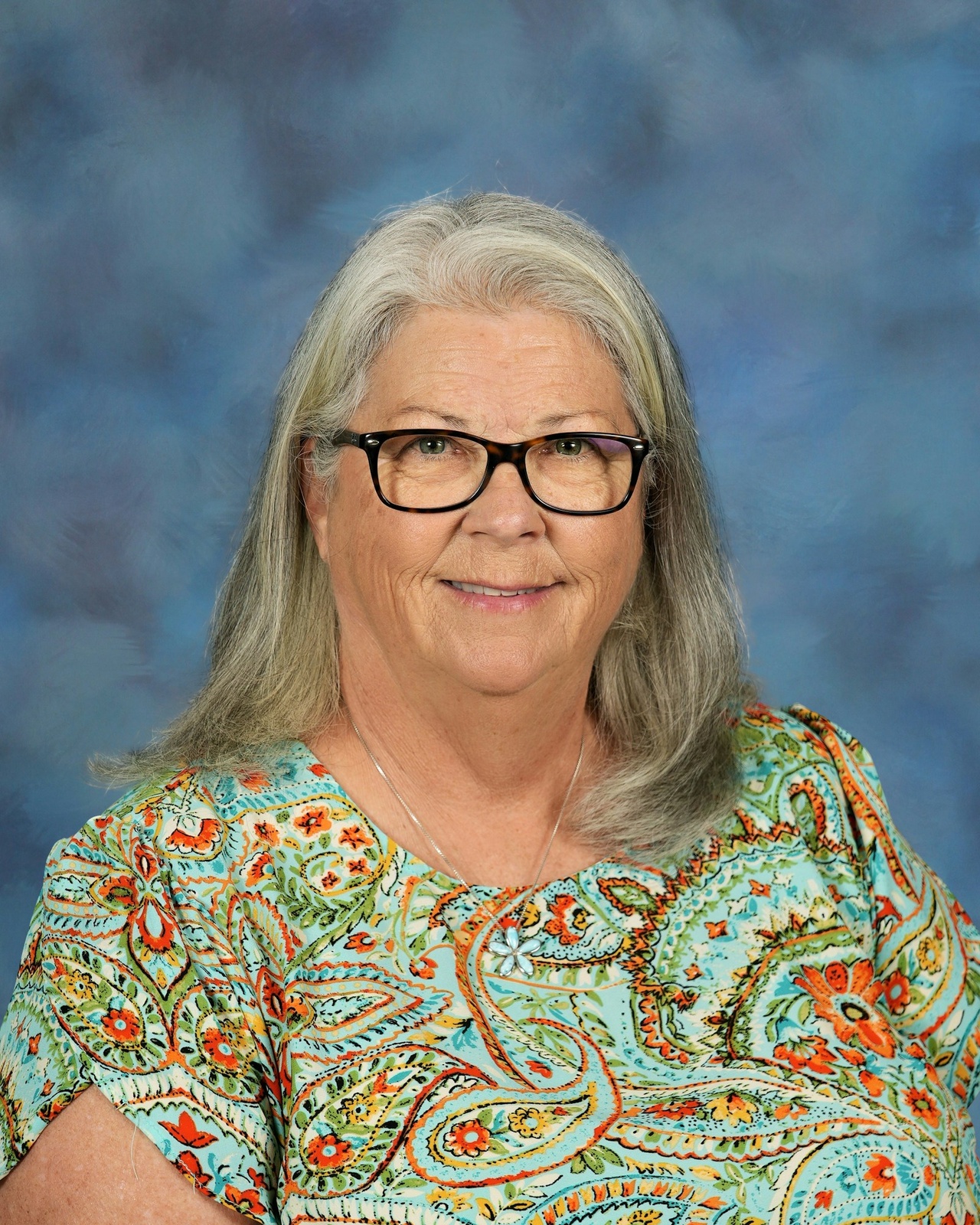 Elderly woman with gray hair and glasses smiling softly. She's wearing a colorful paisley-patterned blouse. Background is a blurred blue gradient.